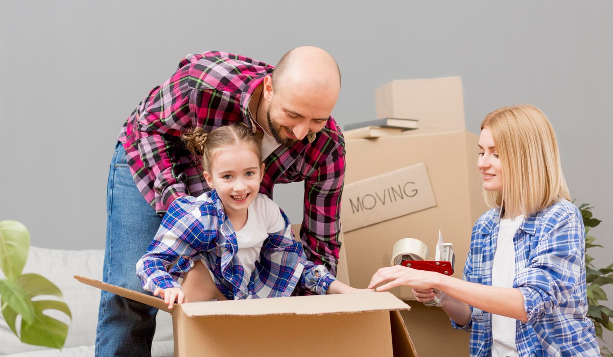 Happy family unpacking boxes together while moving into a new home. A smiling young girl sits inside a cardboard box as her parents pack and organize belongings around her