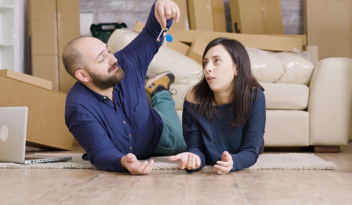 Young couple lying on the floor of their new apartment surrounded by moving boxes and furniture while discussing house keys.