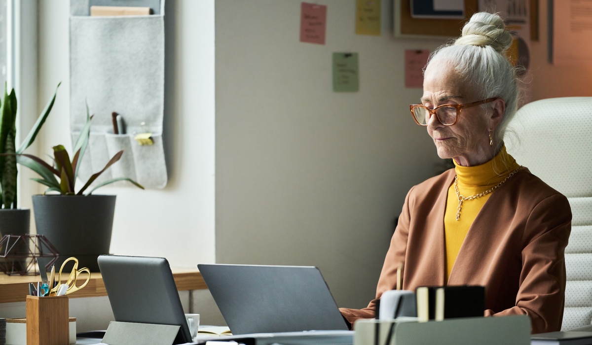 Senior businesswoman working on a laptop in a modern office. She sits at a desk surrounded by office supplies and plants, focused on her screen