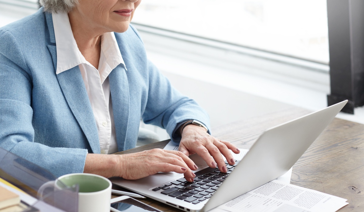 Senior businesswoman typing on a laptop at a wooden desk near a bright window. The image highlights remote work, professional productivity, online administration, or financial management in a modern office or home workspace.