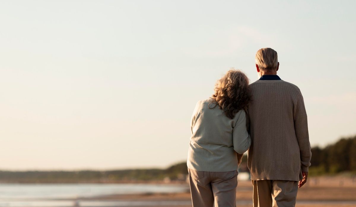 Senior couple walking arm in arm along a quiet beach at sunset. Viewed from behind, the scene conveys companionship, retirement, relaxation, and peaceful aging together by the seaside.
