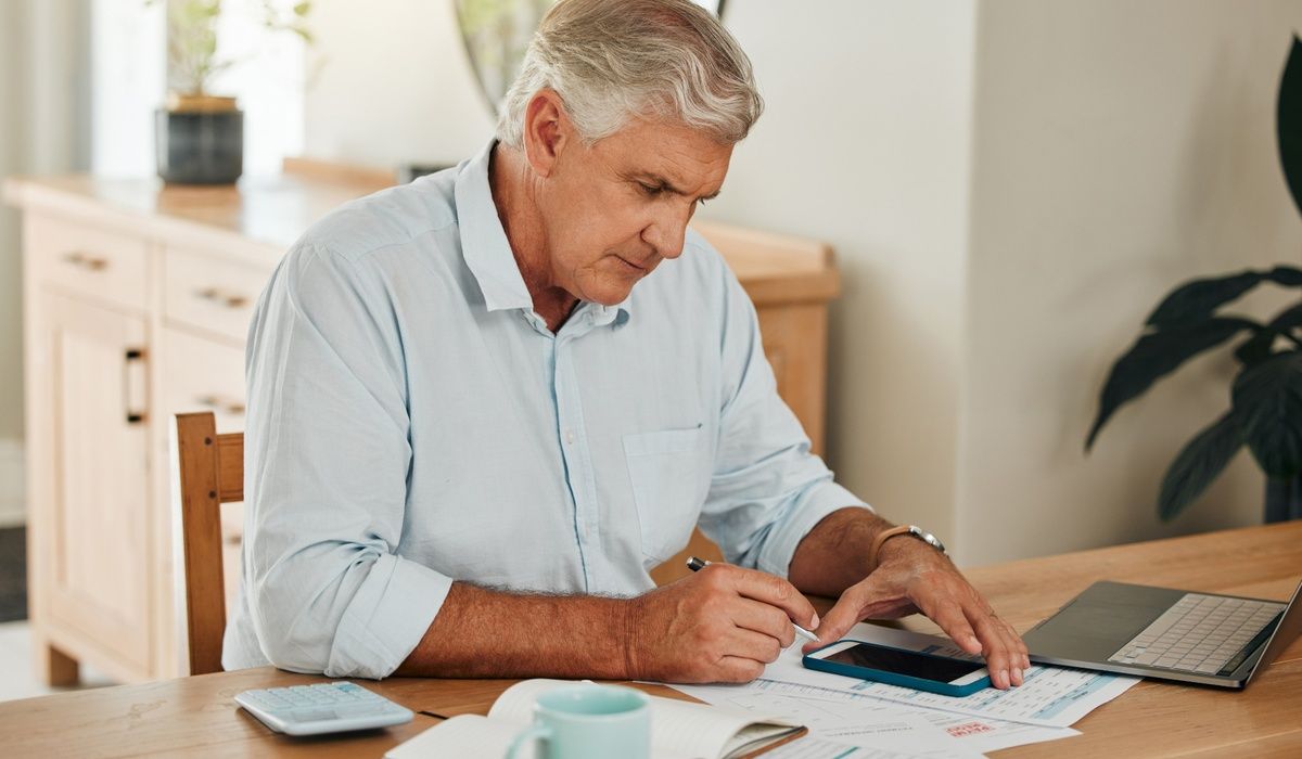 Senior man reviewing financial documents at a dining table while using a smartphone and laptop.