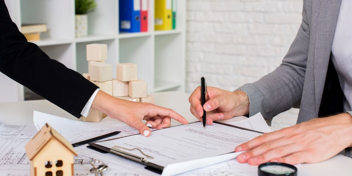 Close-up of two professionals reviewing and signing a contract at a desk with real estate paperwork, a miniature wooden house model, and office supplies.