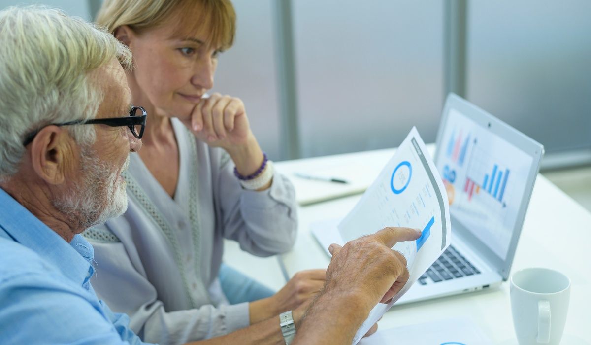 Senior couple reviewing financial reports together at a desk while looking at charts on a laptop.