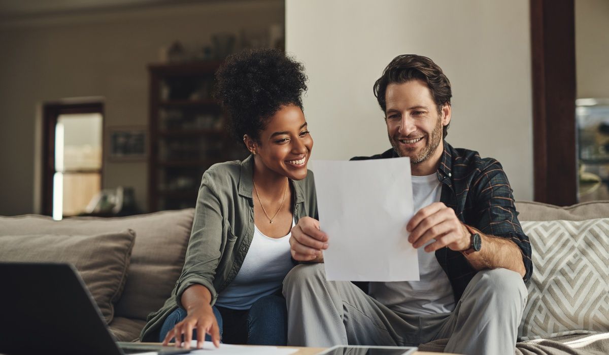 Smiling couple sitting on a sofa at home while reviewing paperwork together on a laptop.