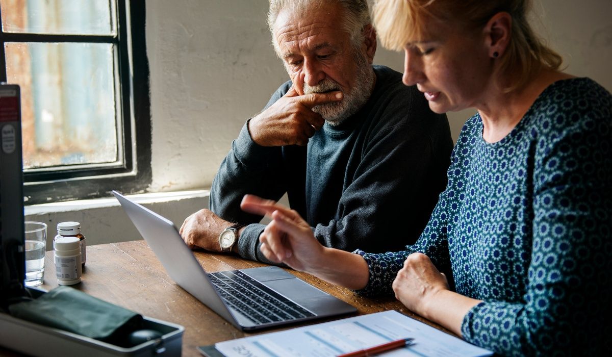 Older couple reviewing information together on a laptop at a wooden table near a window.