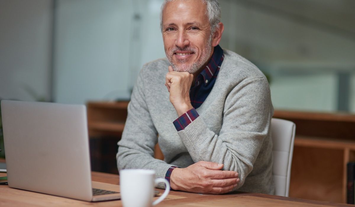 Smiling senior man sitting at a desk beside a laptop and coffee mug in a home office setting. He looks confidently at the camera, conveying professionalism, remote work, online consulting, or successful retirement lifestyle.