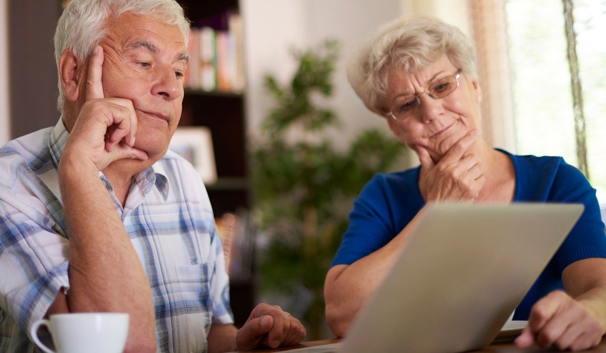 Senior couple looking thoughtfully at a laptop screen while sitting at a table at home. Both appear focused and concerned, suggesting they are reviewing financial information