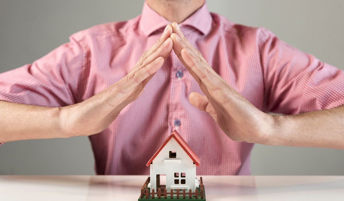 Close-up concept image of home protection and property insurance showing a man forming a roof shape with his hands above a miniature house model.